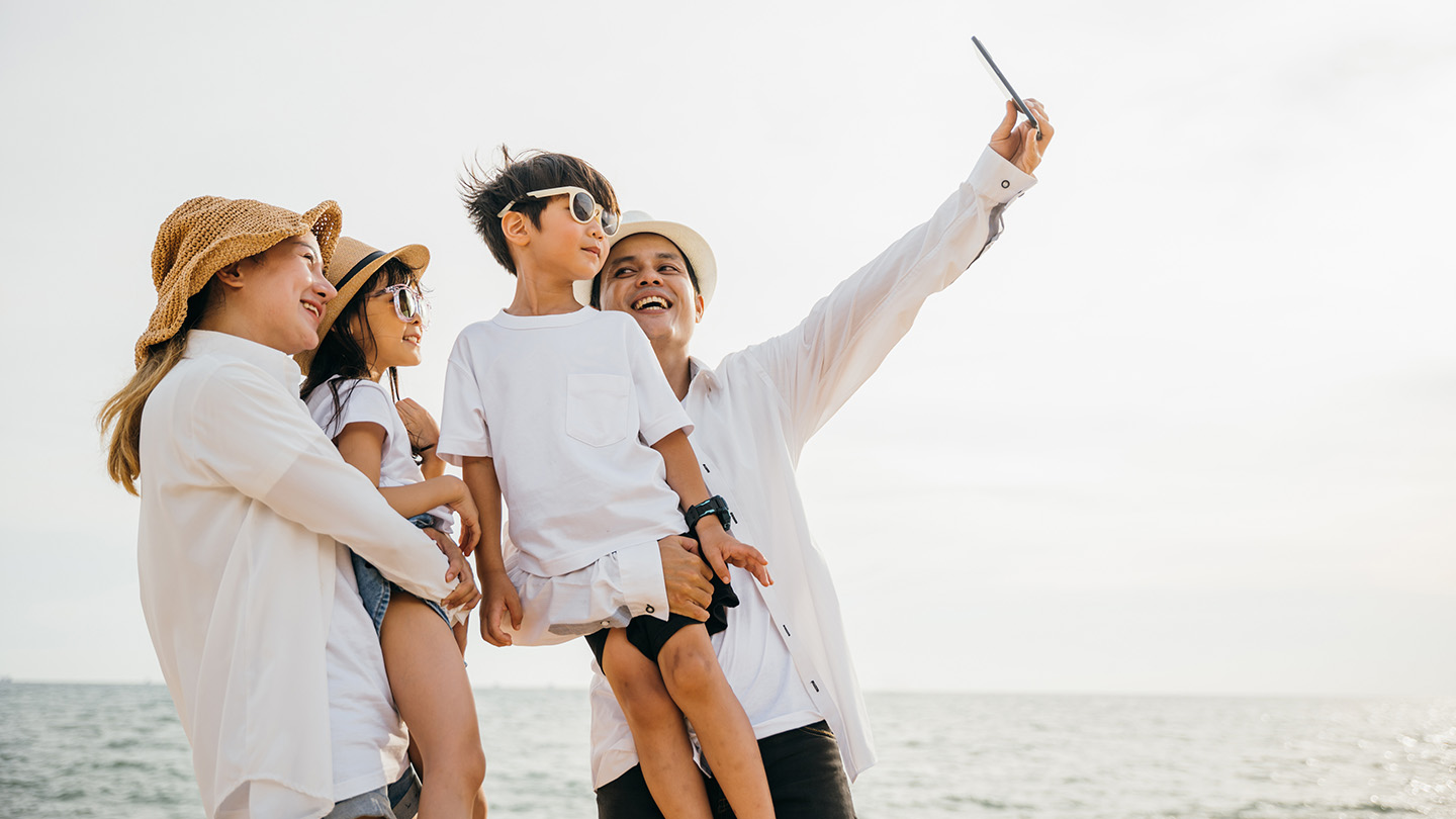 Beachside family fun as they take a cheerful selfie together near the sea. Laughter and joy unfold creating a playful and memorable moment of togetherness during a summer travel adventure.